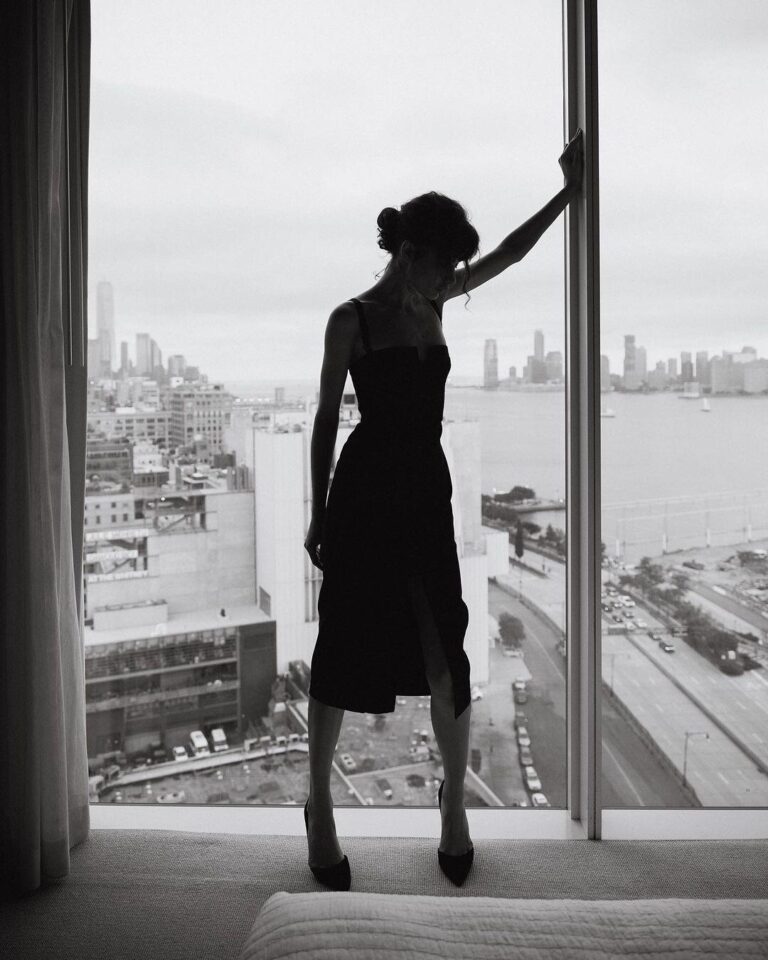 Frau in schwarzem Kleid am Fenster mit Blick über Stadt und Wasser